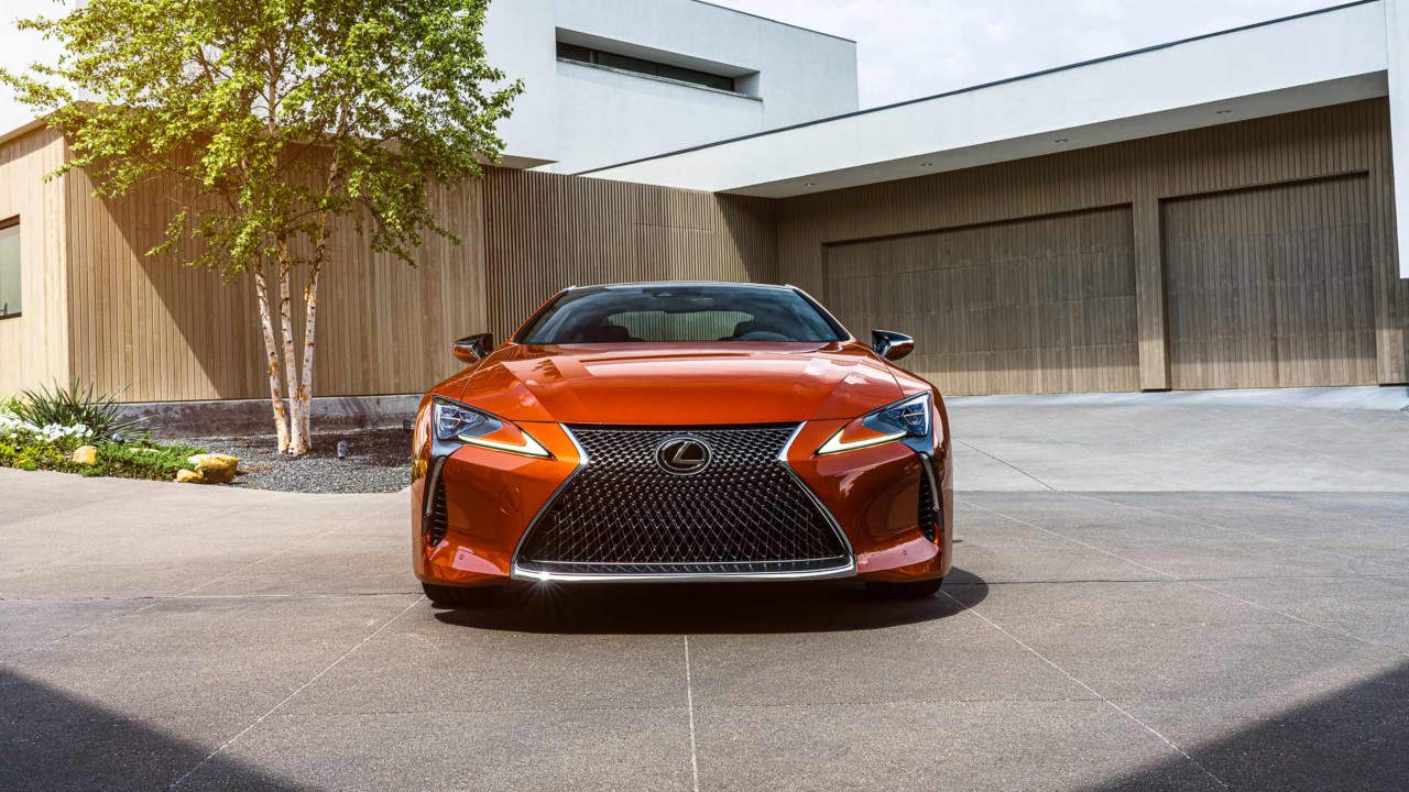 Fordere Ansicht eines orangenen Lexus LC Coupé vor einem modernen Wohnhaus.