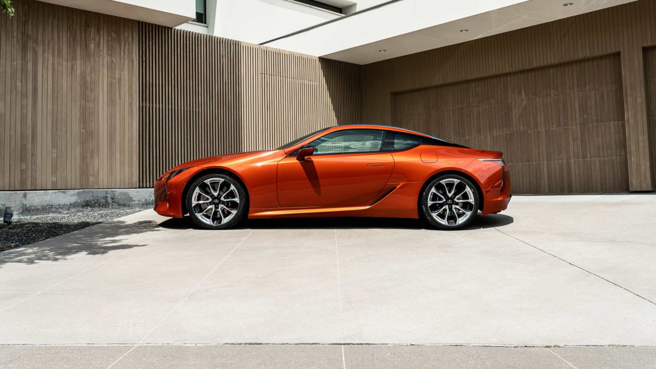 Seitenansicht eines orangenen Lexus LC Coupé vor einem modernen Wohnhaus.